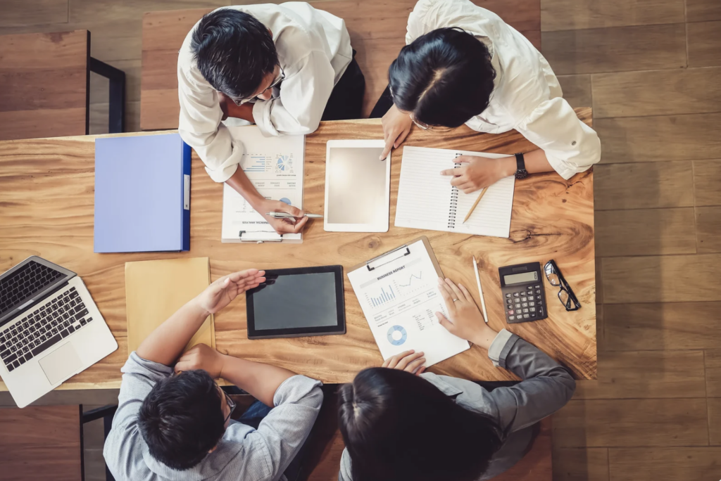 Top view of a team of professionals collaborating on a marketing strategy. The image shows a group of people working together at a wooden desk, reviewing documents, graphs, and charts with a laptop and tablet in use. The scene highlights teamwork, data analysis, and business planning in a modern workspace.